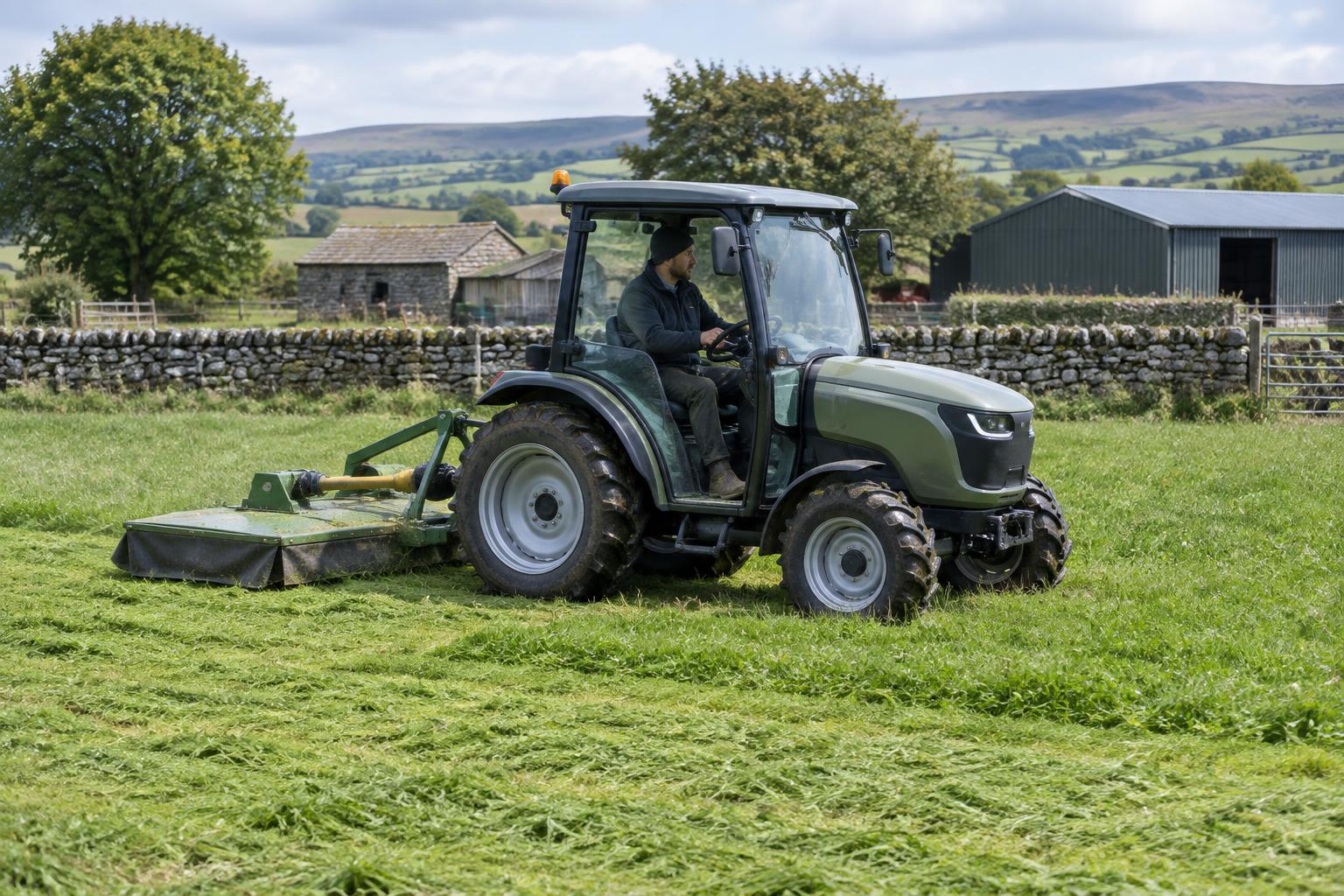Compact electric tractor mowing a grassy paddock beside farm buildings